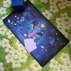 A photo of a blue and purple ghost themed playmat laying on a green and white daisy carpet. The photo is staged with a blue deck box, damage counter dice and sleeved cards. 