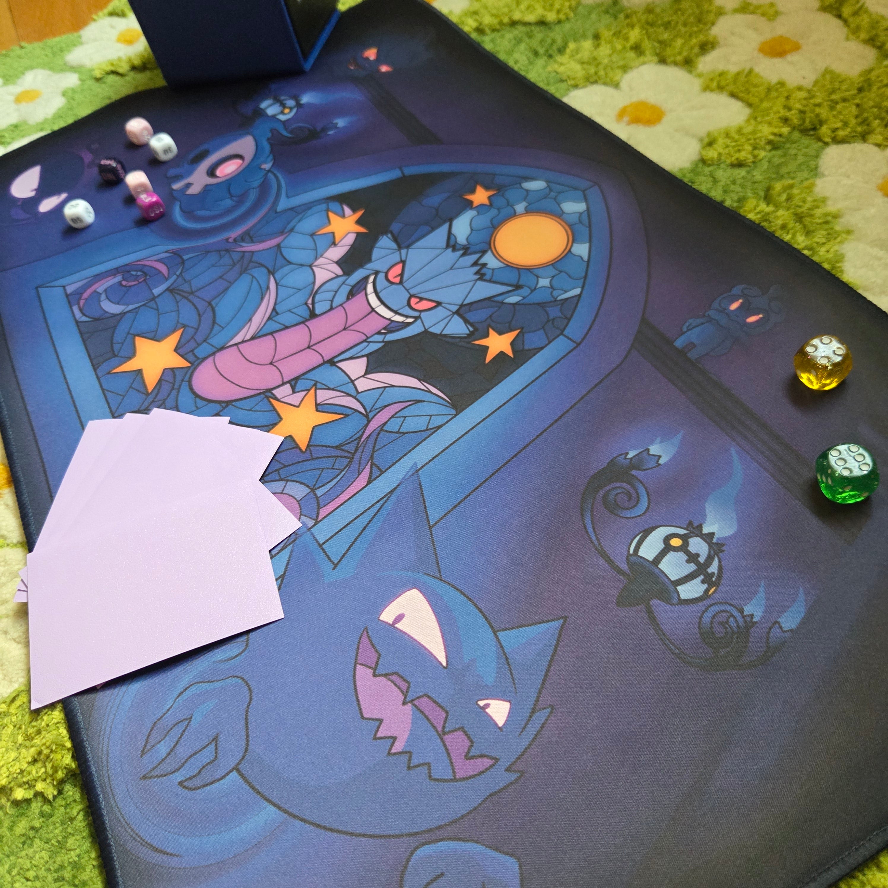 A photo of a blue and purple ghost themed playmat laying on a green and white daisy carpet. The photo is staged with a blue deck box, damage counter dice and sleeved cards. 