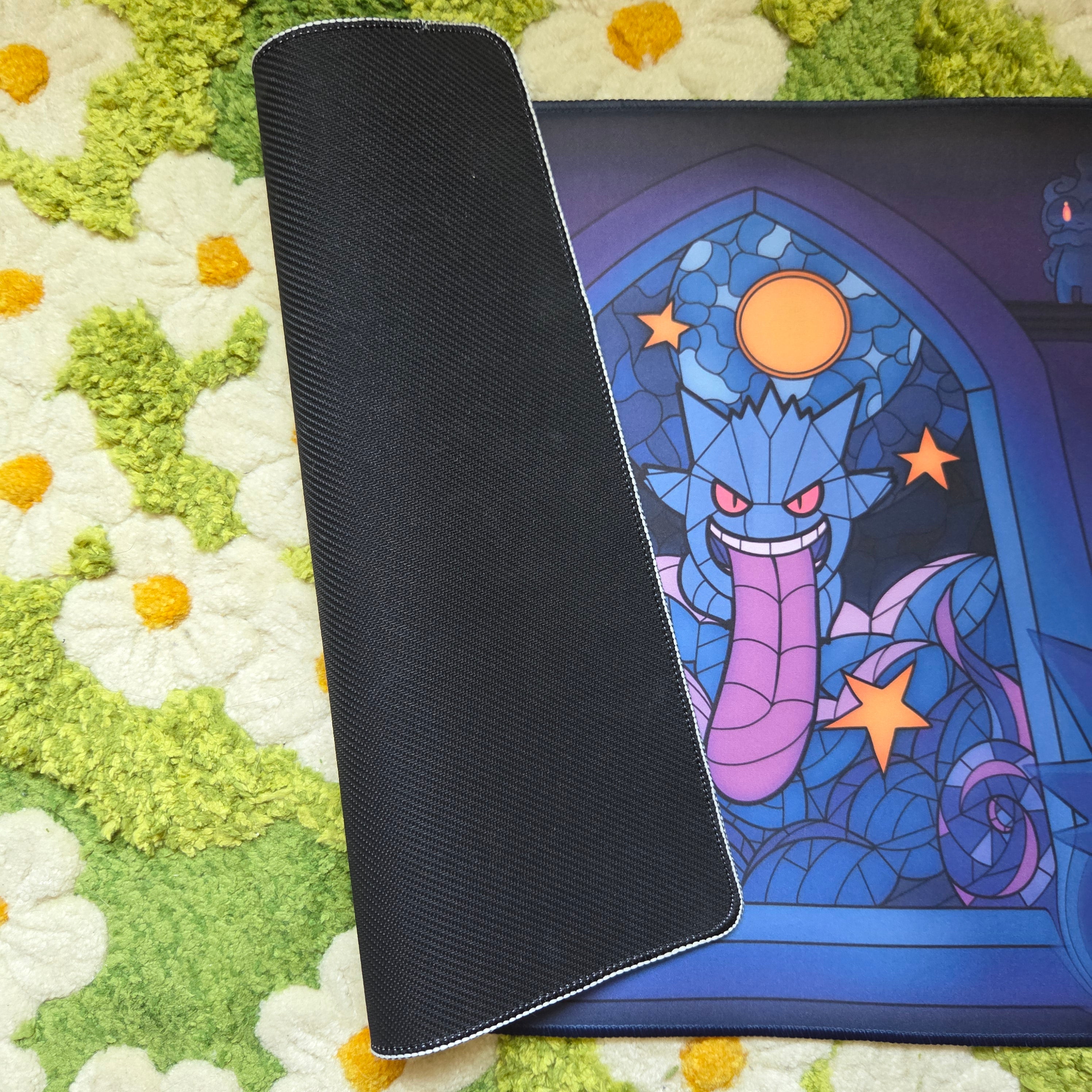 A photo of a blue and purple ghost themed playmat laying on a green and white daisy carpet. The edge of the mat is folded over to display a black textured rubber underside, and white edge stitching.