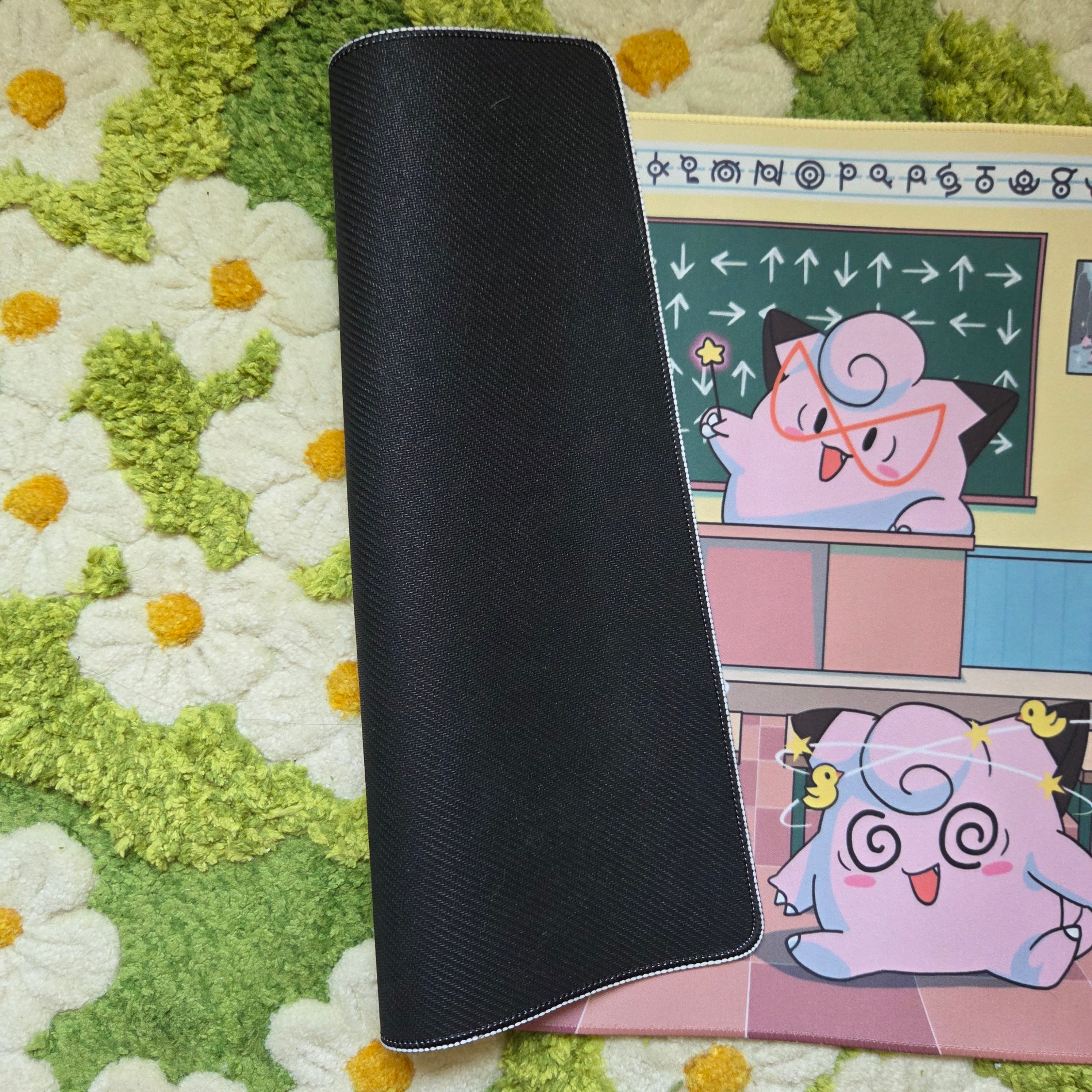 A photo of a yellow, blue, and pink classroom themed playmat laying on a green and white daisy carpet. The edge of the mat is flipped over to display a black textured rubber underside.