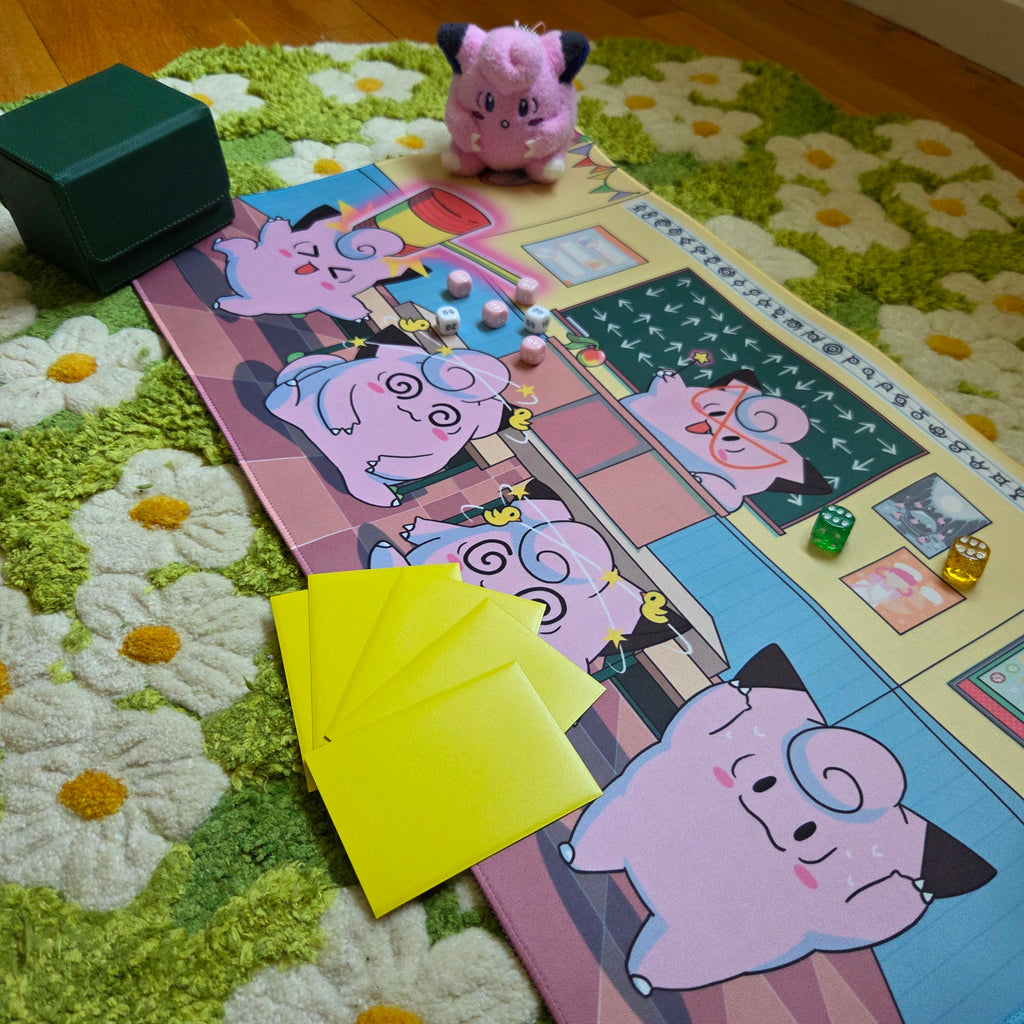 A photo of a yellow, blue, and pink classroom themed playmat laying on a green and white daisy carpet. The photo is staged with a green deck box, a pink plushie, damage counter dice and sleeved cards. 