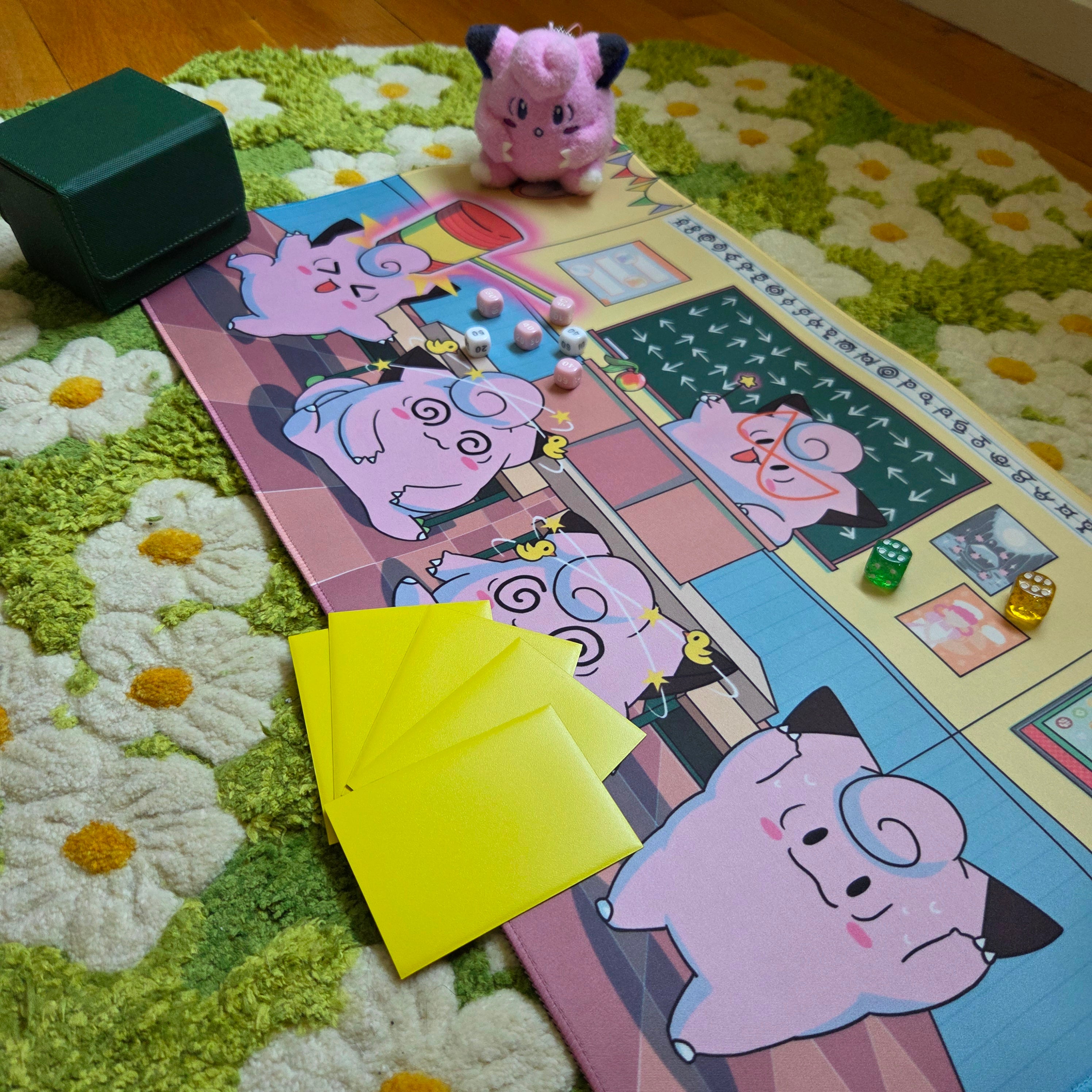 A photo of a yellow, blue, and pink classroom themed playmat laying on a green and white daisy carpet. The photo is staged with a green deck box, a pink plushie, damage counter dice and sleeved cards. 